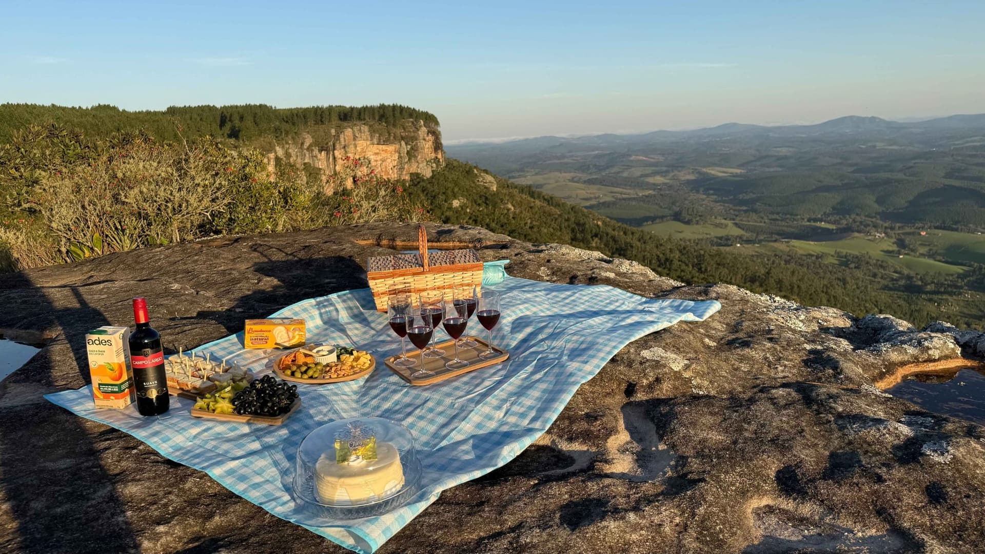 Piquenique ao pôr do sol no alto da Santa do Paredão, com toalha xadrez azul, cesta de vime, taças de vinho tinto, tábuas de frios e frutas e um bolo, diante do paredão de arenito e de um vale com florestas e serras ao fundo, em Jaguariaíva, Paraná.