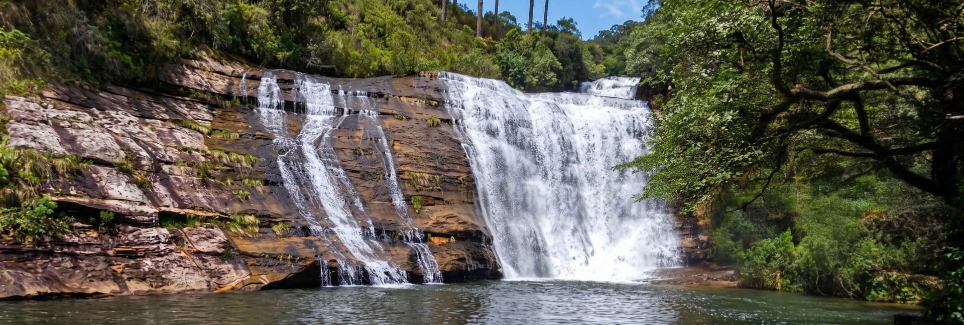 Cachoeira larga de águas brancas caindo sobre paredão de arenito alaranjado, com mata de pinheiros no topo, nos Campos Gerais, Paraná