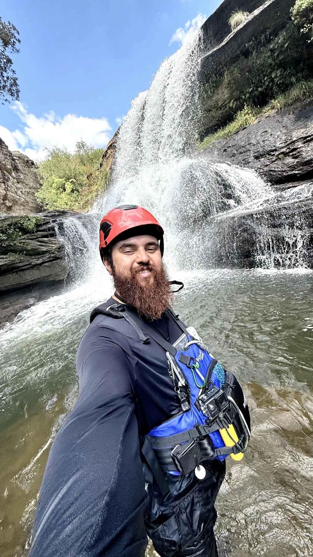 Alisson, guia de turismo da Outventure Ecoturismo, sorrindo de capacete vermelho e colete salva-vidas em frente a uma cachoeira nos Campos Gerais, Paraná